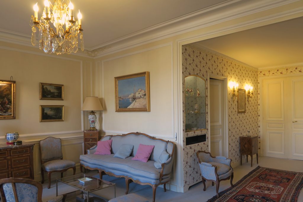 DECOR PEINT : dans un salon décor classique composé de patine spaltée ton paille, avec moulures blanc cassé et rehauts bleus; le ton de fond des vitrines a été choisi avec le tissu, pour mettre en valeur les objets.